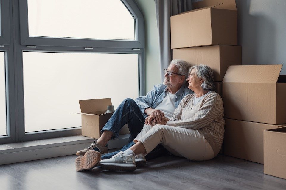 Older couple sitting together beside moving boxes in a bright home, representing downsizing, relocating or planning a renovation.